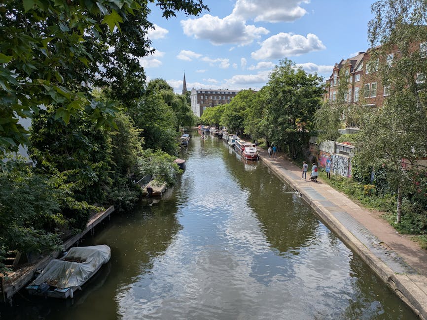 A daytime view of the Lee Navigation canal in Hackney, with calm water reflecting the partly cloudy sky above. On the left side of the canal, there are overhanging green trees with dense foliage, partially covering small wooden docks and a covered boat moored close to the bank. The right side features a paved walkway running parallel to the water, with a few pedestrians walking along it; some are pushing carts or carrying bags. Parked cars line the street adjacent to the walkway, and behind the cars are multistory residential buildings with brick facades, some featuring graffiti on exterior walls. In the background, the canal extends into the distance, bordered by more trees and urban structures, with a church steeple rising above the skyline under a partly cloudy sky. The scene depicts a peaceful urban waterside environment, typical of a home relocation or furniture transport setting near residential areas, with elements associated with moving logistics visible in the surroundings as seen in the image for Man and a Van Hackney's removals services.