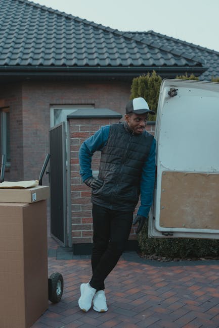 A man dressed in a blue long-sleeve shirt, black padded vest, black trousers, and white sneakers stands outside a residential property in daytime, with his hands on his hips, next to an open white moving van. The van is parked on a brick-paved driveway in front of a house with a grey tiled roof and brick walls, partially obscured by the vehicle. Several cardboard boxes are stacked nearby, some on a trolley with black wheels. The scene depicts the home relocation process, with the man likely preparing to load or unload items as part of furniture transport and packing activities. The environment is well-lit, with natural daylight illuminating the scene, highlighting the equipment used in the moving process, such as the van, boxes, and trolley. The image reflects a professional moving service by '[COMPANY_NAME]', focusing on efficient packing and transportation in the context of house removals in Hackney.