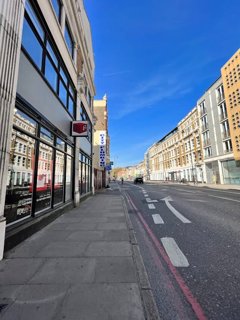 A clear daytime street scene showing the exterior of a building with large black-framed windows and a red postal service box mounted on the wall. The sidewalk is paved with grey concrete slabs, with a single pedestrian walking in the distance. On the right side, a modern multi-storey office building with large windows runs along the street, while further down, a convoy of vehicles is visible. The street features white road markings, including a dashed line and an arrow indicating a turn. The sky is bright blue with no visible clouds. The environment suggests an urban area suitable for house removals or furniture transport, with Man and a Van Hackney likely involved in moving services associated with this location, supporting efficient packing and loading processes for house or office relocations.