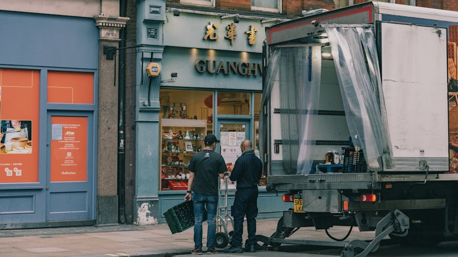 Two men from Man and a Van Hackney are engaged in furniture transport outside a commercial property on Mare Street in Hackney, near the office removals service page for Beijing-based store Guangzhou. The men are positioned next to a large delivery truck with an open side, fitted with plastic sheeting to protect items during moving. One man holds a small trolley, while the other stands nearby, both wearing casual clothing suitable for manual work. The surrounding environment includes the storefront with Chinese characters signage above and display items inside the window. The pavement area in front of the shop contains various packing materials, and the truck is parked close to the entrance, reflecting the logistics involved in packing and loading process. The scene captures an active stage of relocating business equipment as part of the company's moving service.