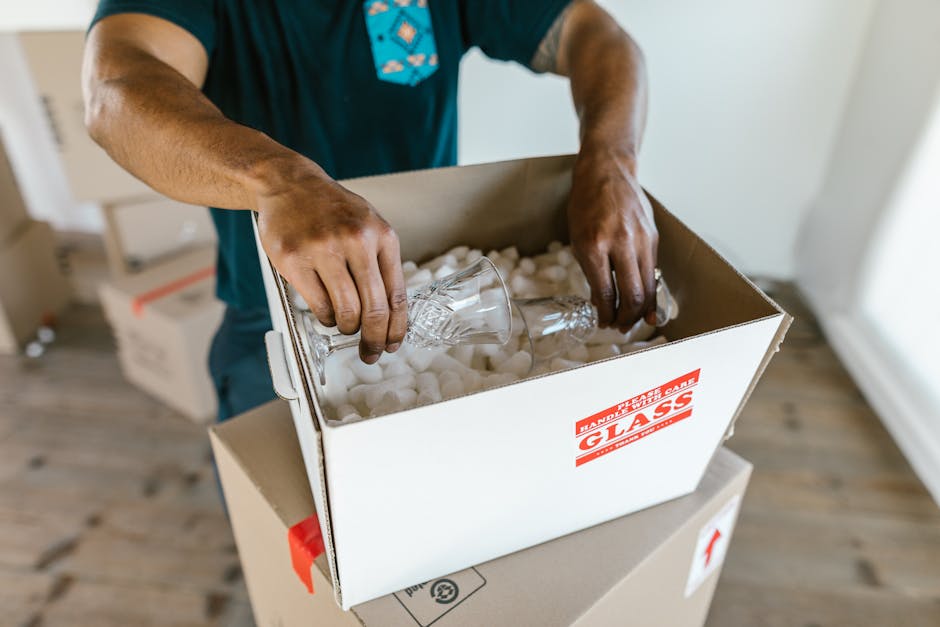 A person wearing a dark t-shirt with a graphic design is seen packing a fragile item, specifically a glass with intricate cut patterns, into a cardboard box filled with white packing peanuts. The box, labeled with red 'FRAGILE HANDLE WITH CARE' and 'GLASS' stickers, is positioned on a flat surface inside a property. Another unopened cardboard box is visible underneath, possibly used for further packing or stacking. The scene is well-lit, with natural light coming from the right side, illuminating the hands, the packaging materials, and the glass. The background includes a wooden floor and a white wall, indicating an indoor environment typical for home relocation or packing and moving services. This process is part of a professional moves coordination by [COMPANY_NAME], focused on careful handling of sensitive items during house removals, with a strong emphasis on packing and loading procedures for safe transport to a new residence.