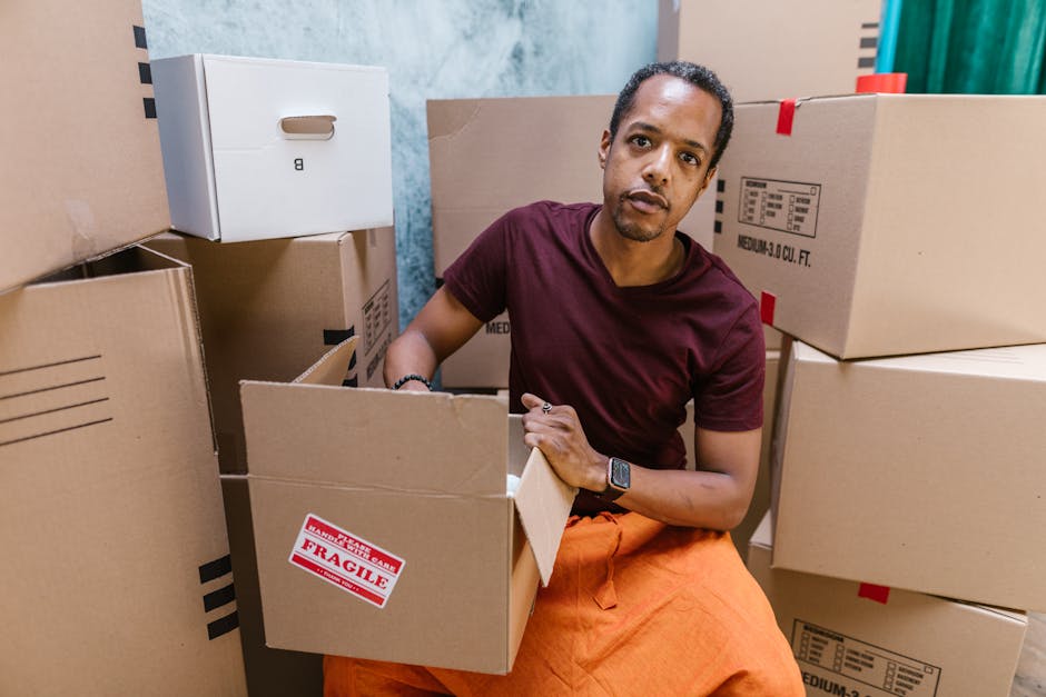A man in a maroon T-shirt and orange pants is sitting among several cardboard boxes in an indoor space, preparing boxes for moving. The boxes are of various sizes, some sealed with packing tape, with one prominently labeled 'FRAGILE' in red. The man is holding an open box in his lap, with his right hand resting on its edge, and is wearing a smartwatch on his left wrist. Surrounding him are packed boxes stacked on top of each other, a few boxes are partially open, indicating the packing process involved in house relocations. The environment features plain walls, with the background showing additional boxes and some visible protective materials like plastic wrap. This scene illustrates part of the furniture transport and packing tasks undertaken by Man and a Van Hackney as part of their house removal and moving services, specifically relevant to professional packing, fragile item handling, and loading procedures for home relocation, in alignment with their Hackney Wick specialist service page.