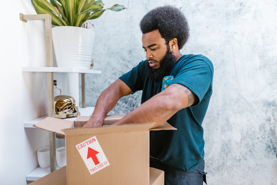 A person wearing a dark t-shirt with a graphic design is seen packing a fragile item, specifically a glass with intricate cut patterns, into a cardboard box filled with white packing peanuts. The box, labeled with red 'FRAGILE HANDLE WITH CARE' and 'GLASS' stickers, is positioned on a flat surface inside a property. Another unopened cardboard box is visible underneath, possibly used for further packing or stacking. The scene is well-lit, with natural light coming from the right side, illuminating the hands, the packaging materials, and the glass. The background includes a wooden floor and a white wall, indicating an indoor environment typical for home relocation or packing and moving services. This process is part of a professional moves coordination by [COMPANY_NAME], focused on careful handling of sensitive items during house removals, with a strong emphasis on packing and loading procedures for safe transport to a new residence.