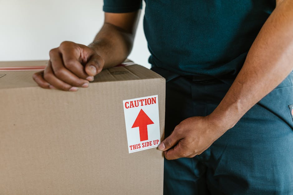 A person wearing a dark green T-shirt and dark trousers is seen holding a large rectangular cardboard moving box, which has a white and red caution sticker on the side indicating 'CAUTION THIS SIDE UP' with an upward arrow. The individual is gripping the box firmly, with one hand on the top edge and the other on the side, preparing for a home relocation or furniture transport. The box is inside an apartment or house environment, positioned on a smooth surface, likely a floor. In the background, a partially visible doorway or hallway suggests the process of loading or unloading during a packing and moving operation. This scene illustrates the careful handling of packed belongings, typical during professional removals provided by companies like Man and a Van Hackney, which specialise in house removals and logistics in areas like Hackney Central E8.