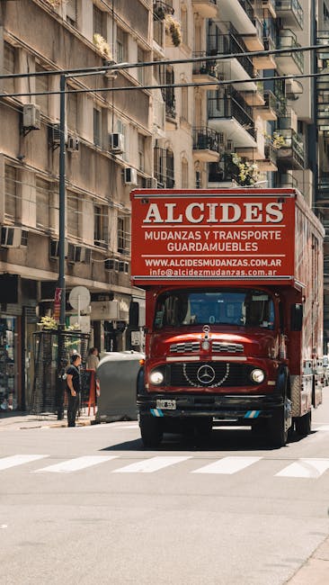A red moving truck with the branding 'ALCIDES' on its large upper sign, positioned on a city street in Hackney with multi-storey residential buildings in the background. The truck is a classic Mercedes-Benz model, and its cargo area is partially visible. Nearby, a person is standing on the pavement, and a few pedestrians are walking along the sidewalk. The scene depicts the process of home relocation, where furniture and packing materials are likely being loaded or unloaded as part of a professional removals service by Man and a Van Hackney. The street environment includes typical urban features such as shopfronts, balconies with plants, and parked vehicles, illustrating the logistics of moving household items in a densely populated area.