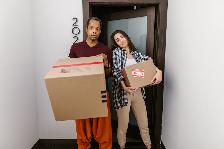 A person wearing a dark green T-shirt and dark trousers is seen holding a large rectangular cardboard moving box, which has a white and red caution sticker on the side indicating 'CAUTION THIS SIDE UP' with an upward arrow. The individual is gripping the box firmly, with one hand on the top edge and the other on the side, preparing for a home relocation or furniture transport. The box is inside an apartment or house environment, positioned on a smooth surface, likely a floor. In the background, a partially visible doorway or hallway suggests the process of loading or unloading during a packing and moving operation. This scene illustrates the careful handling of packed belongings, typical during professional removals provided by companies like Man and a Van Hackney, which specialise in house removals and logistics in areas like Hackney Central E8.