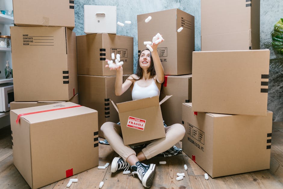 A young woman with long brown hair, seated on a wooden floor amidst numerous large, sealed cardboard boxes, some marked with red 'fragile' labels, in an interior room with light blue walls and a green potted plant visible on the right. She is wearing a white sleeveless top, beige pants, and striped sneakers, and is smiling while catching off-white packing peanuts falling from above. The boxes are stacked at different heights, with some placed on the floor and others on top of each other, indicating a packing or home relocation process. The woman holds a packing peanut in each hand, and a cardboard box in front of her is partially open, revealing more packing materials inside. There is a doorway visible behind her, leading to another room. The scene suggests an organized packing phase of a home removal service, with a focus on careful handling of household items using protective packaging materials, provided by [COMPANY_NAME] as part of their move logistics.