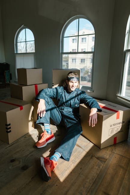 A person in casual clothing and a headband sits on a wooden floor inside a room with large arched windows, surrounded by multiple cardboard boxes sealed with red and black tape, likely prepared for a house move. The boxes vary in size and are placed on the floor around the individual, some leaning against the wall. Natural light from the windows illuminates the space, highlighting the packaging materials and the person's relaxed posture. The room has neutral walls and minimal furniture, suggesting it is part of an ongoing home relocation. The scene depicts a loading or packing process involved in furniture transport and packing for a house removal, with a focus on the logistics of moving typical household items. Man and a Van Hackney is involved in the process, demonstrating the typical stages of packing and preparing belongings for transport during a same-day move in Dalston N16, aligned with the services offered by the company.