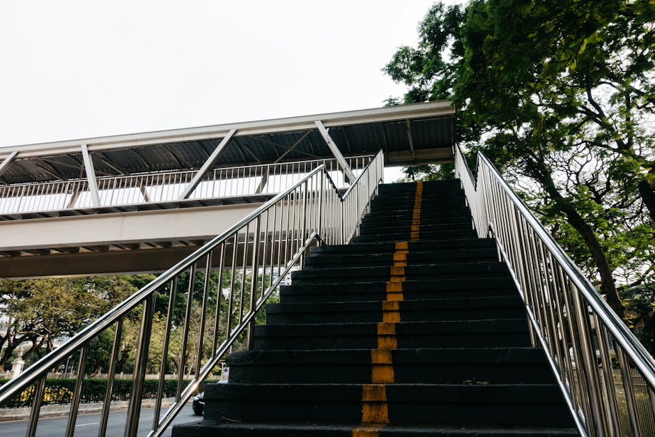 A wide outdoor staircase leading up to a building, composed of multiple concrete steps that appear worn and slightly stained, with metal handrails on each side featuring a geometric pattern. Behind th
