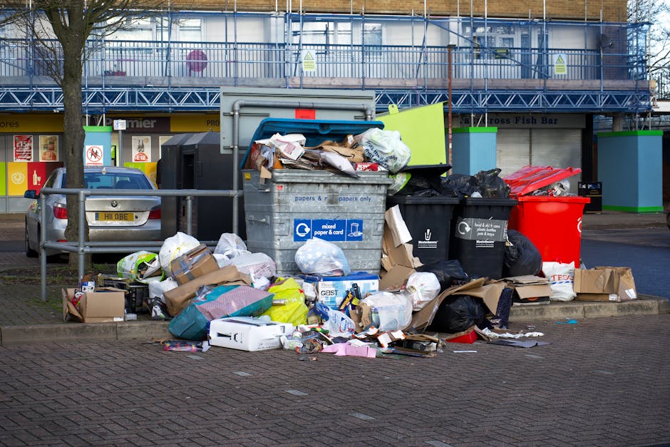 An outdoor scene of overflowing waste and recycling bins positioned on a paved sidewalk in front of a commercial building with ladder scaffolding and windows in Hackney. Multiple black, yellow, and re